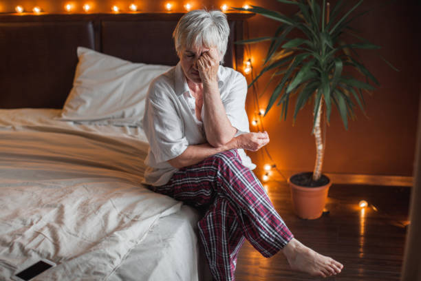 An older woman is sitting on her bed in her pajamas. She has her hand on her face with her eyes closed, as she is stressed out.