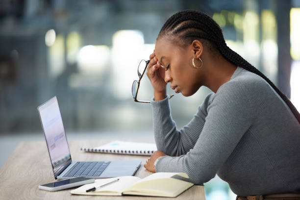 A young African American woman is sitting in front of her laptop with her eyes closed. She has her hand on her head as she looks stressed out.