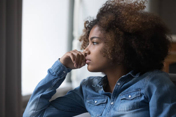 A teenage African American girl is sitting in her home with a distressed look on her face. She is leaning her head on her hand.