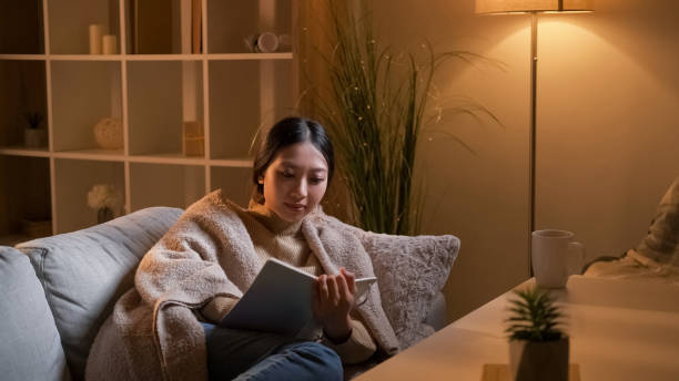 A young Asian American woman is laying on her couch in the living room. She is cuddled up with a blanket and is reading a book.