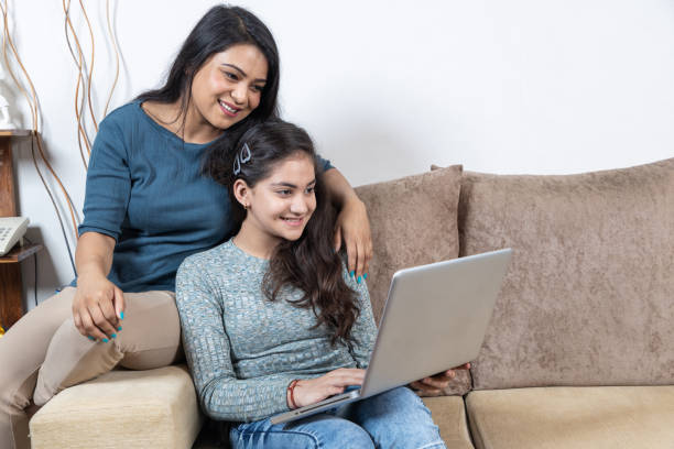 A young girl and her mother are sitting together on the couch as they smile. They are both looking at the open laptop on the teenage girls lap.