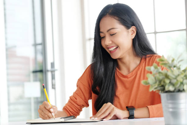 A young Asian American woman is sitting at her office desk working. She is writing in her notebook as she smiles.