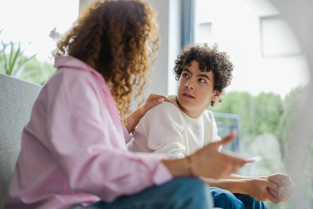 A teenage boy and his mother are sitting beside each other on the couch. They are both looking at each other.