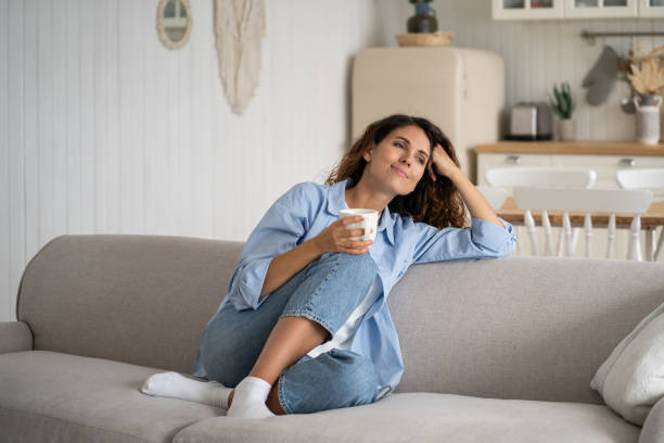 A young woman is sitting in her living room on the couch. She has a cup of coffee in her hand as she smiles.