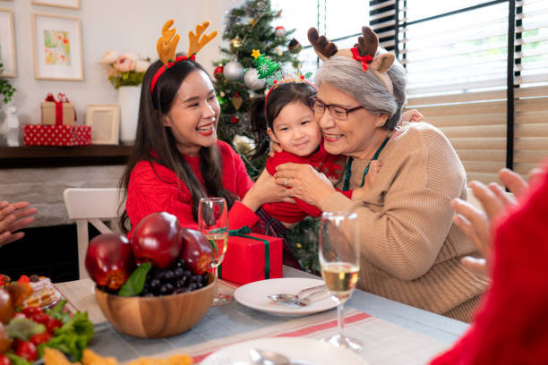 An Asian American mother, daughter, and grandmother are dressed up for the holidays and sitting on the dinner table. They are all hugging as they open gifts.