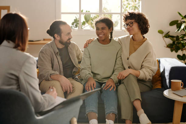 A biracial family of three is sitting across from a therapist in the therapy office. The teenage girl is sitting in between her mother and father as they smile.