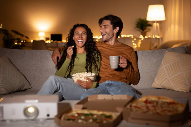 A biracial couple is sitting together on the couch watching a movie. They are eating popcorn and drinking hot chocolate.