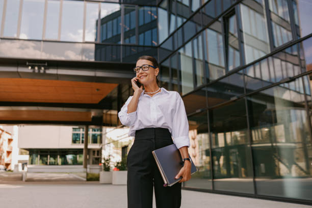 A businesswoman is standing in front of her office building with her notebooks in her hand. She is on a phone call as she holds her phone up to her ear.