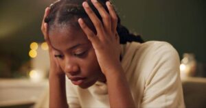 A young African American woman is sitting in her home, with her hands holding her head.