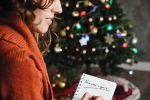 A young woman is writing down her new years goals on a notepad. Behind her is a christmas tree.