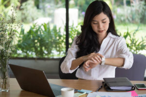 An Asian American business woman is sitting at her desk in her office. She is looking at her watch on her hand. In front of her is an open laptop.