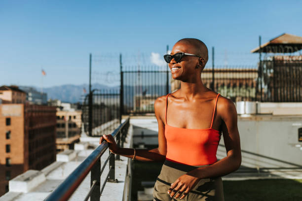 An African American woman is standing on a rooftop looking at the city. She is wearing sunglasses and smiling.