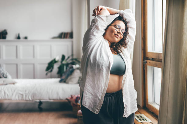 A young biracial woman is in her home stretching her arms above her head. She has her eyes closed with a smile on her face.