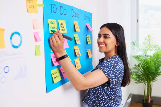 A young woman is adding to her to-do list on her wall. She is smiling as she writes on the sticky note.