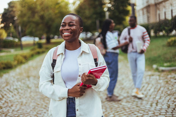 A young African American female student is walking on campus with her backpack on her. She is holding red books in her hand and is smiling.