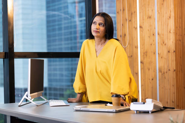 An Indian woman is standing in her office leaning on her desk. She is looking up as she thinks.