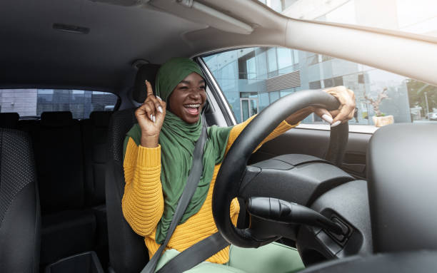 A Muslim African American woman is driving her car, wearing a green hijab. She is singing and has her hand up in the air.