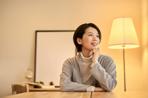 An Asian American woman is sitting on her dining table in her home. She is looking out the window as she smiles.