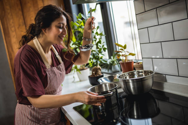 A Hispanic woman is wearing an apron as she cooks over the stove. She is taste testing her soup with a ladle.