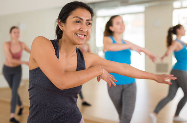 A Hispanic woman is taking an exercise class among 4 other women. She is dancing as she smiles.