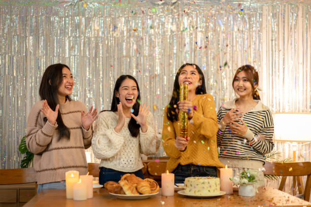 A group of four Asian American women are celebrating a birthday together. One of the women is blowing confetti as the rest of the women clap.