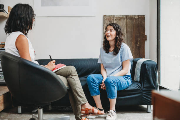 A young woman is sitting across from her therapist in a therapy office. The therapist has a notebook in her lap and a pencil.