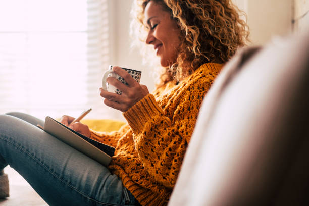 A young woman is sitting on her couch in the living room. She is writing in her journal, as she holds a cup of coffee in her hand.