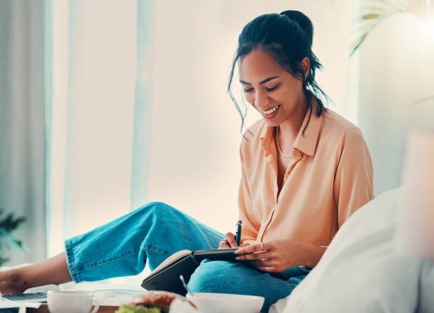 A young biracial woman is sitting on her bed in her bedroom. She is writing in her notepad as she laughs.