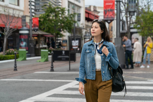 A young Asian American woman is walking in the city across the crossway. She is wearing glasses on her head.