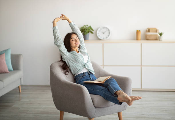 A young woman is sitting in a chair in her office. She is stretching her arms above her head. There is an open notebook on her lap.