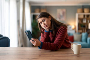 A young woman is sitting in her home at the dinner table. She is looking at her phone with a distressed face.