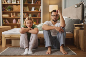 A young couple is upset with each other and sitting beside one another on the floor next to their bed.