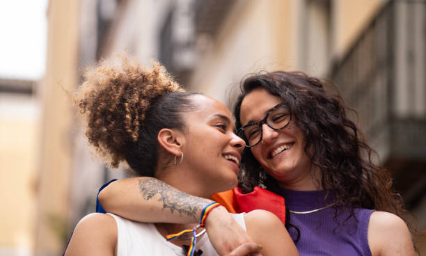 A same-sex lesbian couple is walking together outside. One of the women have their arms around the others as they smile.