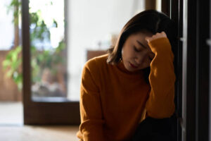 An Asian American woman is sitting in a dark room with her head down and her hand on her forehead.