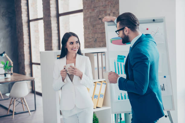 A businessman and businesswoman are standing together in their office as they flirt.