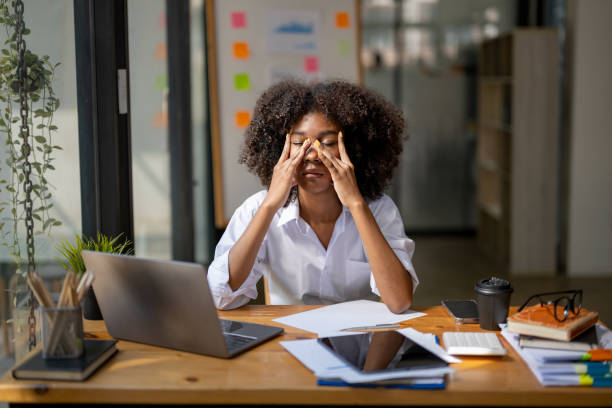 An African American businesswoman is sitting at her desk in her office, stressed out with her eyes closed and her hands on her face.