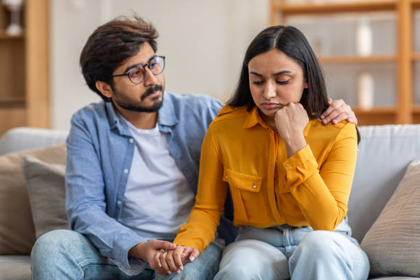 An Indian couple is sitting beside each other on the couch. The man is holding the womans hand and has his arm over her shoulder.