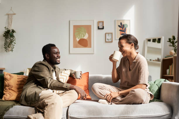 A young African American couple is sitting together on their couch in their home. They are drinking coffee together as they laugh.