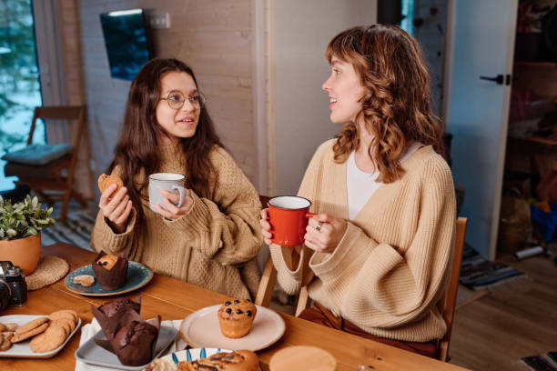 A mother and her daughter are sitting at the dinner table, eating muffins and drinking coffee together.