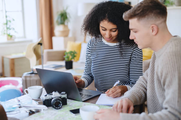 A biracial young couple is sitting together at a desk doing work. The woman is writing on a notepad with her laptop open.