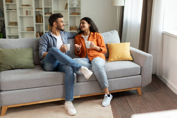 A young Indian couple is sitting beside each other on the couch as they drink coffee.