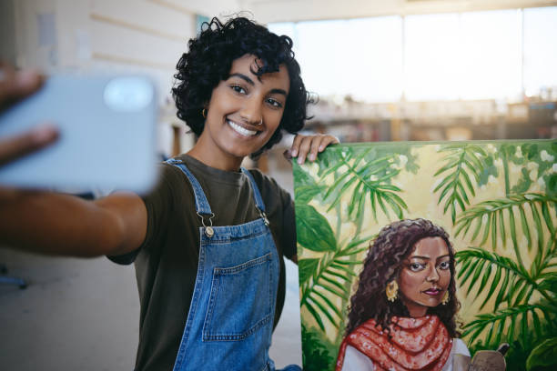 A young Indian woman is standing next to a painting she created, as she takes a selfie with her phone.