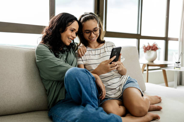 A mother and her daughter are sitting together on the couch looking on the daughters phone.