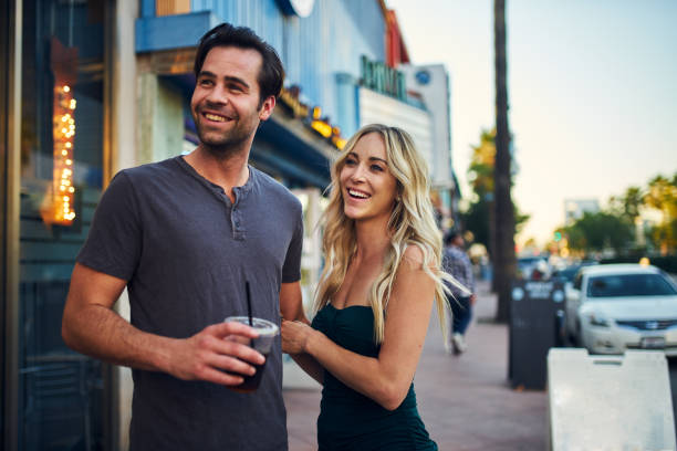 A young couple are standing on the sidewalk together as they smile. The man is holding a cup of coffee in his hand.