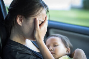 An Asian American mom is sitting in her car covering her face, as her baby sleeps in her arms.