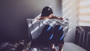 A woman is sitting on her bed in a dark room. She has her pillow on her lap as she covers her face.