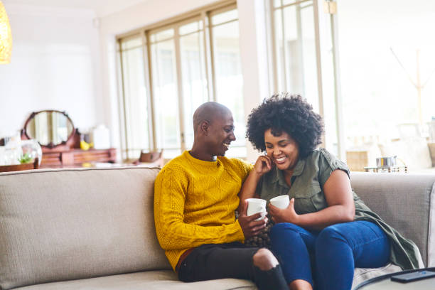 An African American couple is sitting in their home on the couch laughing and drinking coffee.