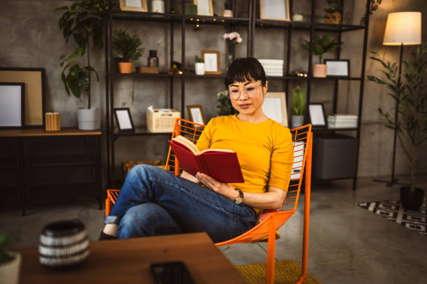 An Asian American woman is sitting in a cafe, reading a book on an orange chair.