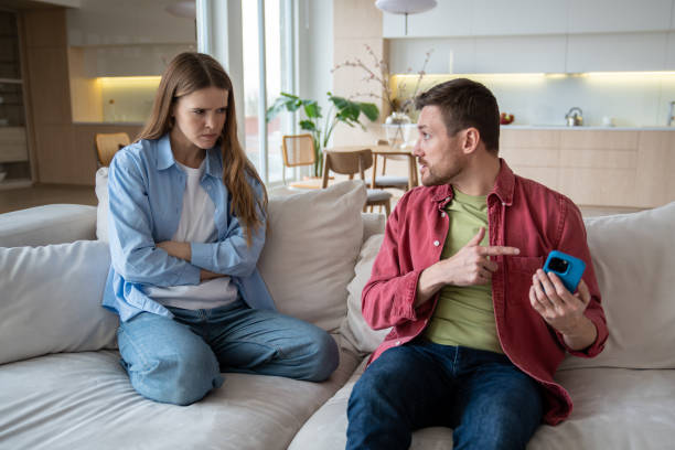 A young couple is sitting on the couch together. The woman is upset with her arms crossed, as her partner points at the phone in his hand.