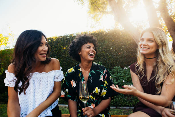 Three female friends are sitting outside having a picnic. They are drinking wine and laughing together.